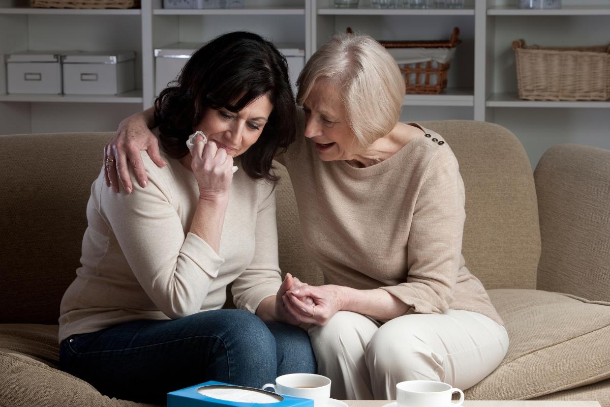 Older woman in beige sweater comforting distressed younger woman on couch, emotional support between family members