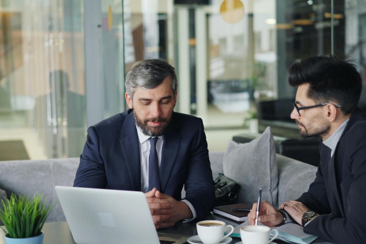 Two businessmen in suits having productive discussion at modern office table with laptop and coffee