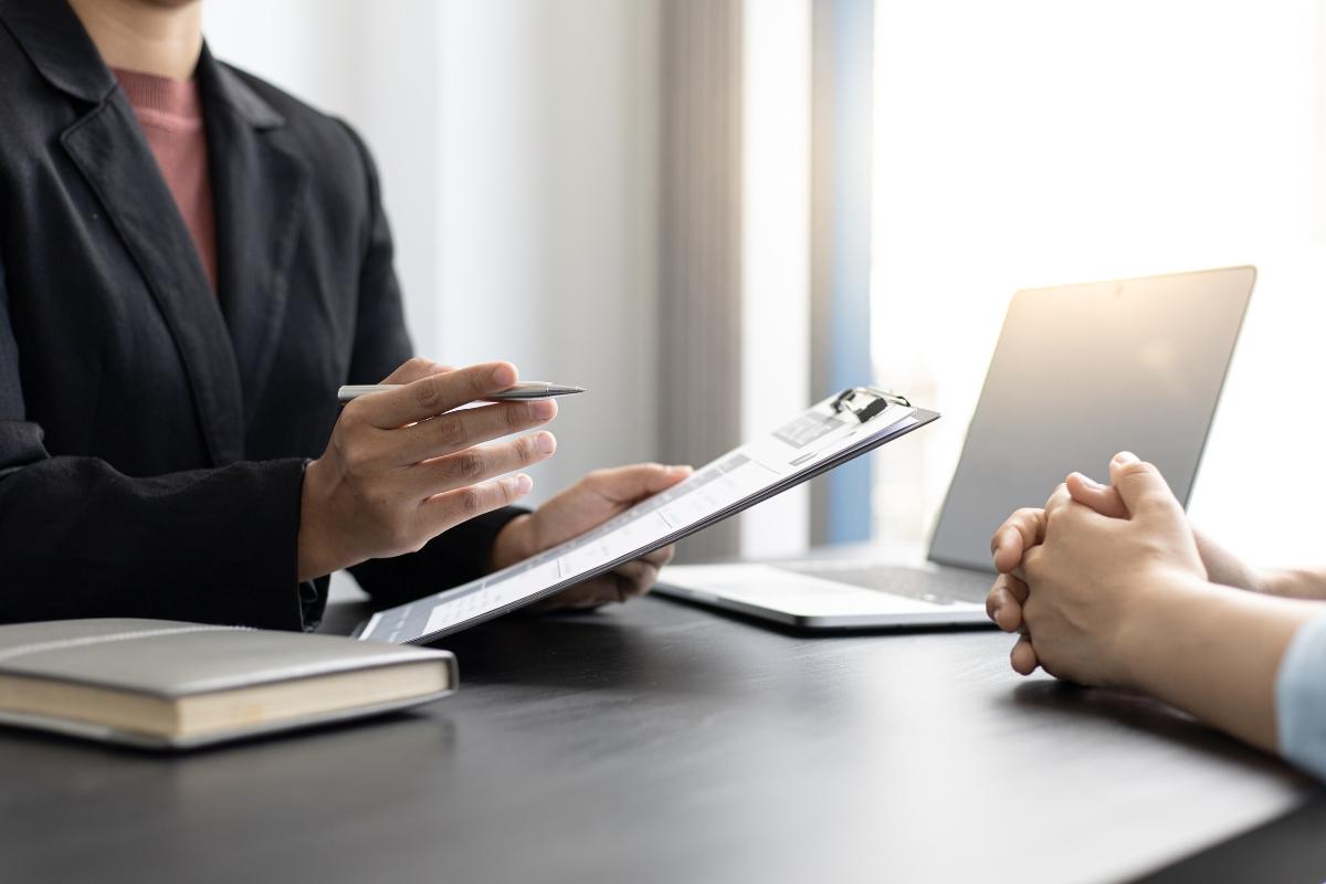 Business professional holding clipboard during consultation meeting across desk from client with laptop