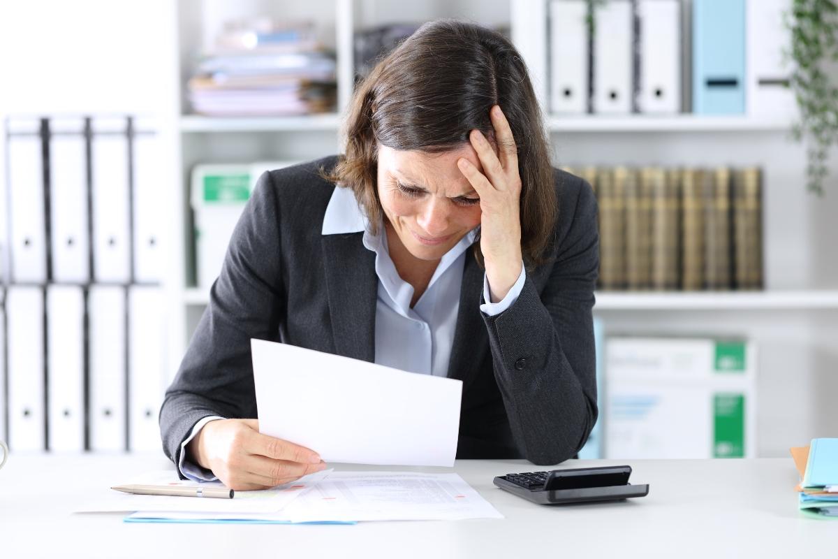 Worried female executive in business attire reading document at desk with hand on forehead showing workplace stress
